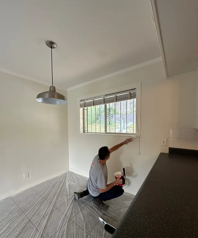 Painter brushing window trim in a dining area with floors covered in plastic
